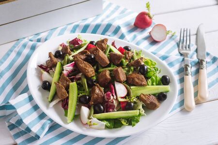 Beef cooked with salad mix, green beans, cucumbers, olives and radishes. On a white plate. On a striped white blue napkin. Fork and knife.の写真素材