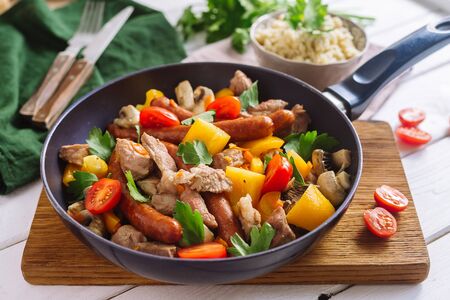 Frying pan with chopped pork meat, yellow bell pepper, cherry tomatoes, sausages, mushrooms, parsley leaves. A frying pan stands on a wooden board, stands for hot. Couscous grits in a bowl.の写真素材