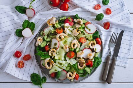 Iceberg salad with chicken fillet rolls pieces, thinly sliced radish, cucumbers, spinach leaves, cherry tomatoes, Chimichurri sauce. Flat lay on a white wooden table, with kitchen towel and utensils.の写真素材