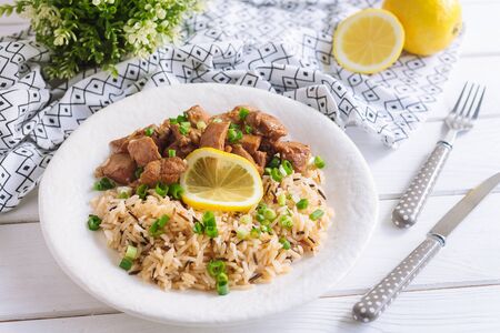 A white plate with steamed wild rice with sliced pork slices, garnished with a slice of lemon. The dish is sprinkled with chopped scallion. On a white wooden table, with a napkin, and utensils.の写真素材