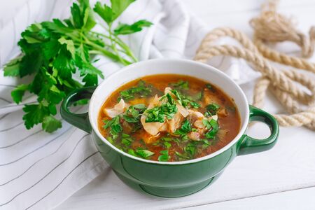 Soup with fish on chicken stock, with celery and tomatoes in a deep green bowl. The dish is sprinkled with chopped parsley leaves. On a white wooden table with a white kitchen towel.の写真素材