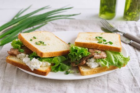 White wheat bread toasted sandwich with pork meat and cheese, salad and sour cream. Chopped chives, bottled olive oil, a knife and a fork. Against the background of a gray fabric napkin.の写真素材