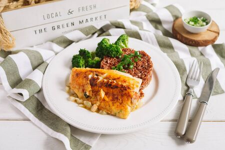 Cod fish fillet on a white plate with brown rice and broccoli. Sliced green onions in a white bowl on a wooden stand. White wood background. Fork and knife. Striped fabric napkin.の写真素材
