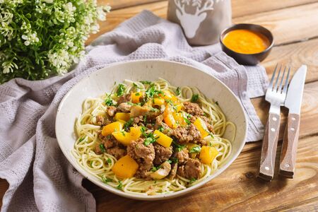 Deep plate with spaghetti, minced meat, chopped parsley, yellow bell pepper. Nearby is a bowl with sauce, a fork and a knife, a table napkin, a kitchen waffle towel. Serving per person.の写真素材