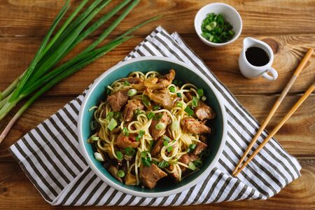 Egg noodles with pork, oyster mushrooms and chili sauce. Chopped green onions, soy sauce, chopsticks. Striped fabric napkin. Wooden table, wooden background. View from above.の写真素材