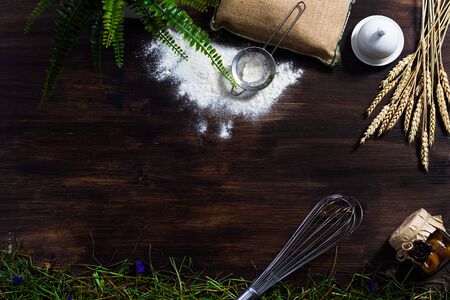 Wooden dark background on which lie a whisk, flour, a bag, a sieve, a jar, a jar of jam, straw, hay, oatmeal ears, sprinkled flour, leaves of fern, canvas bag, cooking utensil, ball whisk.の写真素材