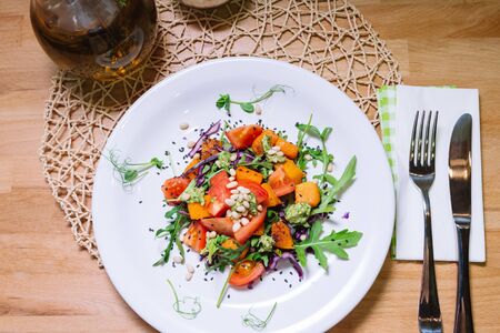 Warm salad with pumpkin, tomatoes, arugula, pesto sauce with pine nuts and black sesame seeds on white plate top viewの写真素材