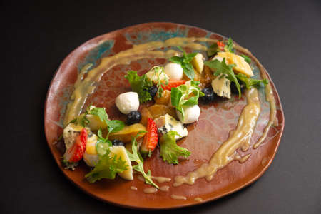Cheese and fruit salad on a handmade plate. Mozzarella, blue cheese, parmesan, zucchini rolls, micro greens, blueberries, strawberry wedges, caramelized pear, pear puree sauce. Side view of a plate.の写真素材