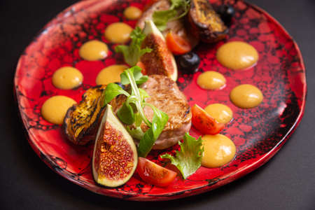 Warm salad, fried pork, fig slices, fried peaches, blueberries, cherry tomato slices, green leaves, pumpkin puree sauce. On a handmade red plate and a dark background.の写真素材