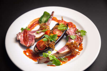 Lamb ribs with baked eggplant, cherry tomato wedges, caramelized onions, small basil leaves, micro greens, with red sauce on a white plate on a dark black background. Side view.の写真素材