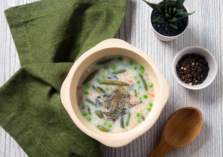 Hot vegetables soup made from green beans, green peas, carrots, basil leaves, with protein broth in a deep beige bowl. Wooden spoon, green linen napkin, black peppercorns, gray wooden backgroundの写真素材