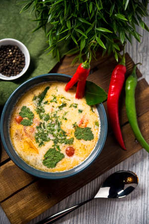 Tom yum soup portion on a wooden board and table. Slightly above view. Decorated with hot pepper and herb leaves. Green table cloth and a spoon on a side.の写真素材