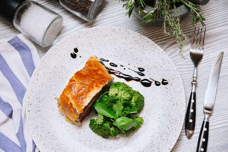 A slice of beef wellington with a portion of steamed broccoli dressed with the thick balsamic sauce on a white plate. Knife and fork on the side. Small cloth of napkin in a corner.の写真素材