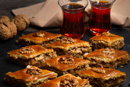 Honey baklava with walnuts on black slate. Portions of oriental sweets. Turkish sugary pieces. Tea poured in armudu glass in the background.の写真素材