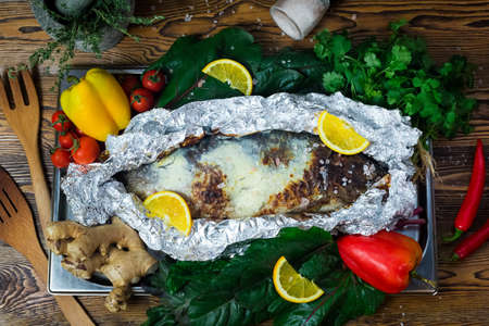 Baked whole fish in foil. The fish is sprinkled with coarse salt. Surrounded with bell peppers, lemon slices, leafy greens, spicesの写真素材