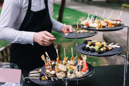 Waiter places plates on the table. catering service. wedding welcome food. Fruits on skewers and...