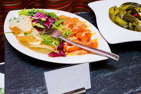 Salmon marinated in sea salt, lettuce leaves, and thin slices of croutons. A plate of pickled sliced cucumbers on another plate. The buffet table.の写真素材
