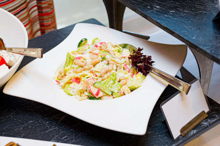 A plate of crab salad on the buffet table. Salad of fresh cucumbers, crab sticks, white beans, iceberg lettuce leaves, Lola rossa. dressed with mayonnaise.の写真素材