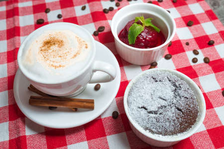 A cup of cappuccino with a portion of chocolate fondant in a ramekin under powdered sugar and raspberry sorbetの写真素材