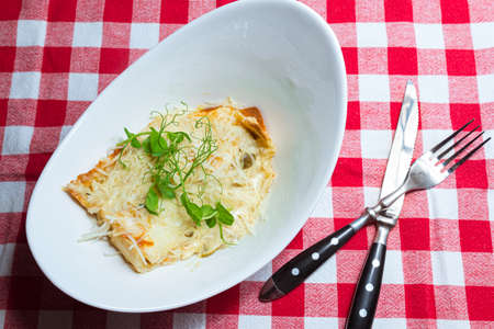 A large portion of homemade lasagna in a deep white plate garnished with micro greens on a checkered red and white napkinの写真素材