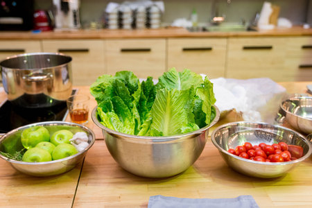 Raw foods and ingredients on the kitchen table before cooking: green apples, lettuce leaves, bowl of cherry tomatoes, casserole on the electric stoveの写真素材