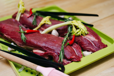 Raw beef fillets seasoned with fresh herbs, garlic, and red chili peppers on a green cutting board, prepared for cooking.の写真素材