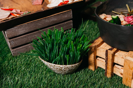 A basket filled with fresh chives on an artificial grass surface with rustic wooden crates and a bowl of vegetables in the background.の写真素材