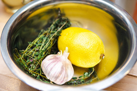 A stainless steel bowl containing fresh ingredients: a whole lemon, garlic bulb, and aromatic herbs, ready for culinary use.の写真素材