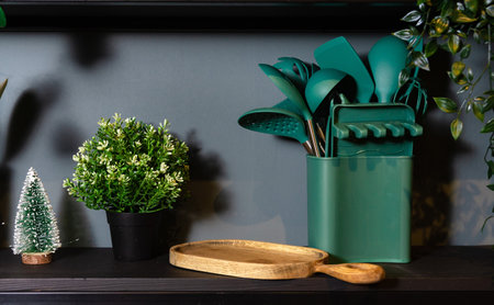 Green kitchen utensils organized in a container next to potted plants and a wooden tray on a dark shelf.の写真素材
