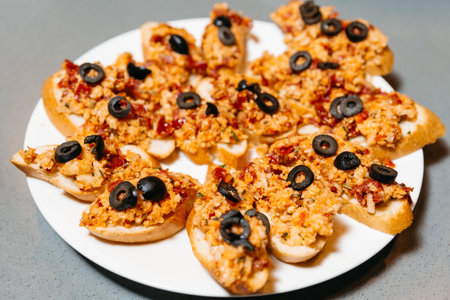 Sliced baguette topped with a flavorful mixture of diced tomatoes, garlic, and black olives, arranged on a white plate.の写真素材