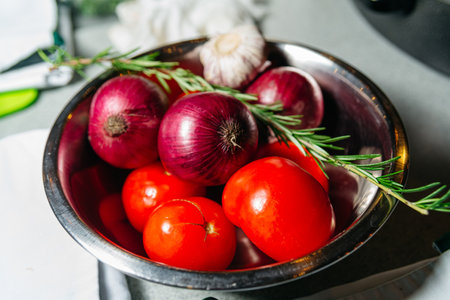 Bright red onions, ripe tomatoes, and fresh rosemary sprigs in a metal bowl, ready for cooking.の写真素材
