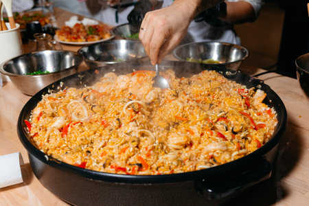 A chef's hand stirs a large pan of seafood paella, rich with rice, shrimp, and mussels, a traditional Spanish dish.の写真素材