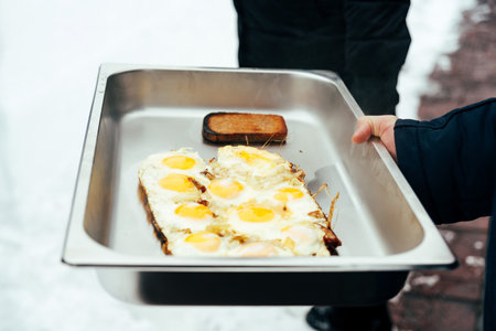 A person holding fried eggs with toast in a large tray outdoors during winter, showcasing a casual camp brunch setup.の写真素材