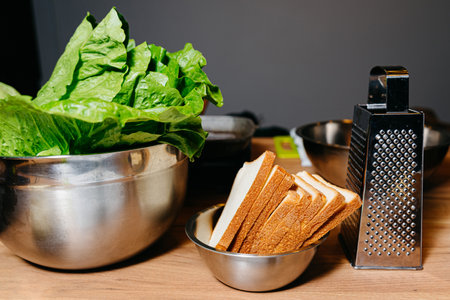 A stainless steel bowl full of crisp green lettuce next to a small bowl of sliced bread and a metal...