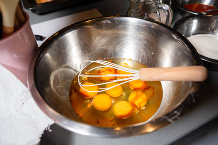 Preparing a meal by whisking fresh eggs in a stainless steel mixing bowl with a wire whisk, with...