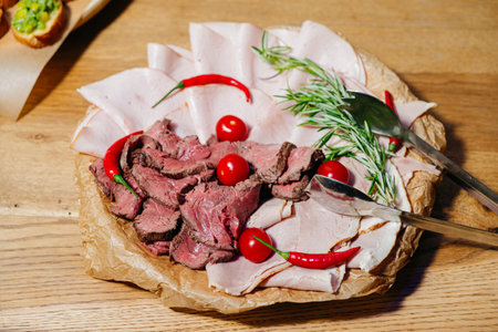 Top view of a charcuterie platter featuring sliced roast beef, ham, cherry tomatoes, chili peppers, and rosemary garnish.の写真素材