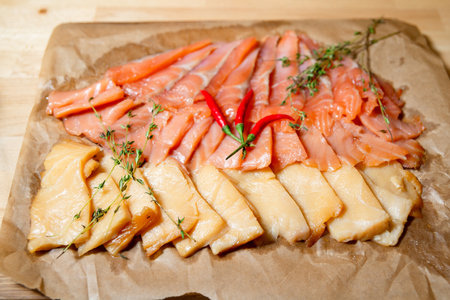 Assorted slices of cold smoked fish arranged on parchment paper, garnished with red chili peppers and sprigs of fresh thyme.の写真素材