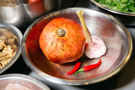 A stainless steel bowl with a whole pomegranate, a head of garlic, and two red chili peppers...