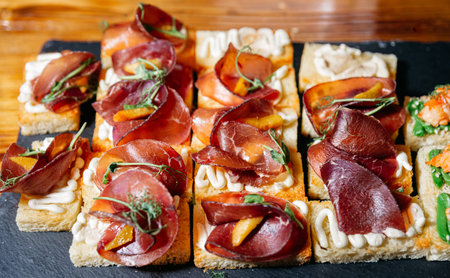 Elegant bruschetta canapÃ©s made with toasted bread slices, topped with whipped cream, cured meat, herbs, and garnish, served on a slate tray.の写真素材