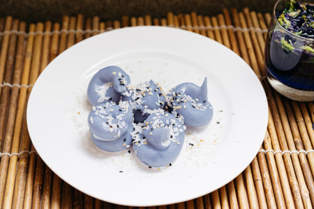 Blue-hued Thai dessert dumplings made from butterfly pea flower, topped with shredded coconut and sesame seeds, served on white plate.の写真素材