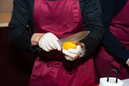 Person in gloves and apron carefully slicing a peeled orange with a kitchen knife during a cooking class or food prep.の写真素材