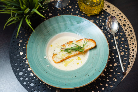 A gourmet bowl of creamy soup garnished with toasted bread, shredded cheese, fresh rosemary, and a drizzle of olive oil.の写真素材