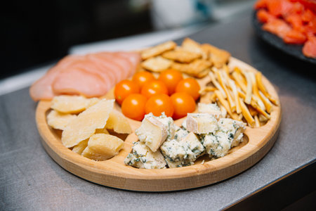 Wooden serving board filled with blue cheese, hard cheese, cherry tomatoes, breadsticks, crackers, and deli meat for appetizers.の写真素材