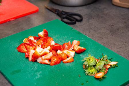 Bright red strawberries cut into quarters on a green cutting board, with stems discarded beside them, ready for dessert prep.の写真素材
