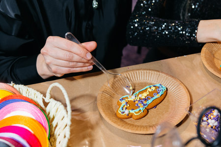 Person using a spoon to decorate a gingerbread cookie with colorful icing and sprinkles during a...