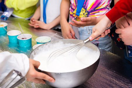 Kids mix foamy liquid in metal bowl using whisk during a fun hands-on cooking science experiment, likely making ice cream.の写真素材