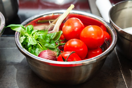 Bowl filled with vibrant red tomatoes, basil leaves, red onion, and garlic, showcasing fresh ingredients for cooking or salad prep.の写真素材