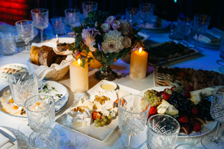 Upscale dining setup with elegant glassware, floral centerpiece, candles, assorted cheeses, bread basket, and fresh fruit for guests.の写真素材