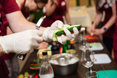 Close-up of gloved hands peeling a fresh cucumber with a peeler in a culinary workshop or cooking class setting.の写真素材
