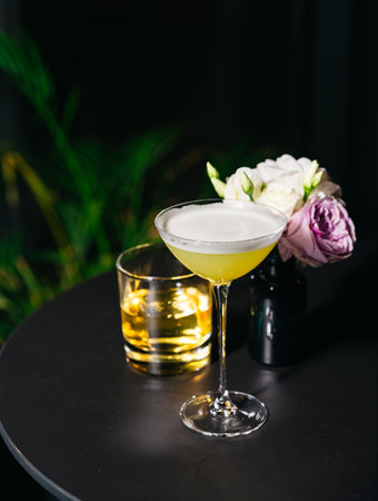 Close-up of a yellow cocktail with foam, served on a dark table next to a whiskey glass and small flower vase.の写真素材
